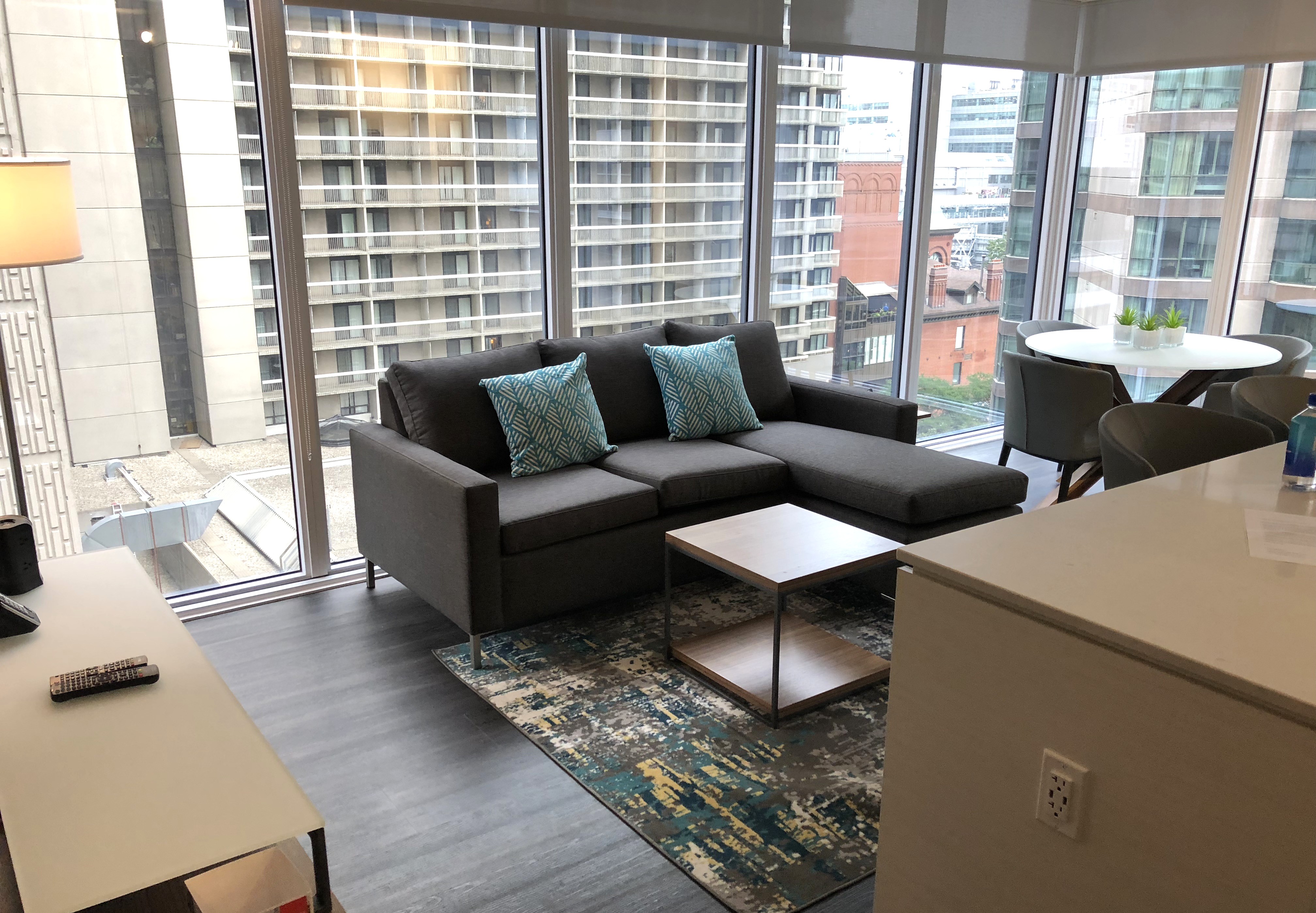 Designer open-concept living area at Livemore featuring a grey sectional sofa with turquoise pillows, a white waterfall kitchen island, and expansive floor-to-ceiling windows with urban views.