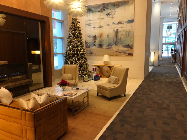 Luxury Furnished Condominiums Toronto: Elegant residential lobby at The Ritz-Carlton featuring a marble coffee table, tan leather armchairs, a large abstract mural, and a festive holiday display.