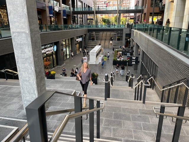 Wide-angle exterior view of the multi-level retail promenade at The Well, featuring modern architectural stairs, Shoppers Drug Mart, and glass-railed walkways.