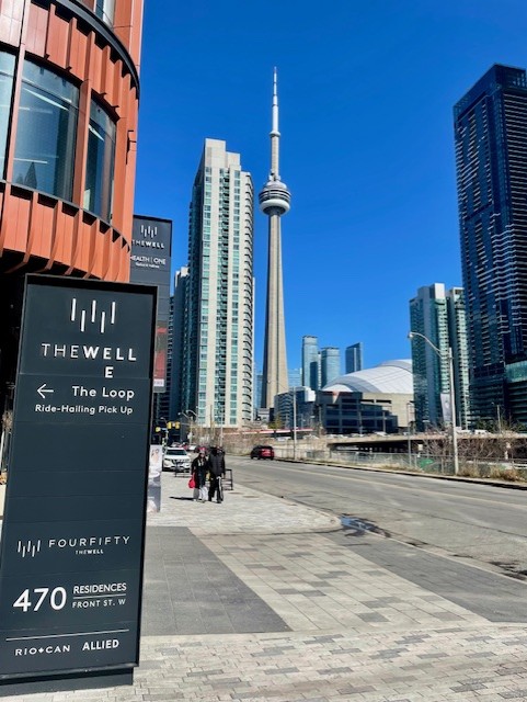 Street-level view of The Well signature signage at 470 Front St W, showing the CN Tower and Rogers Centre in the background under a clear blue sky.